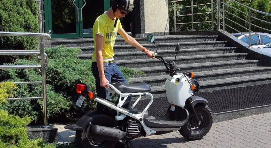 Chłopak w kasku i żółtej koszulce przy białym skuterze. Zastanawiasz się, ile kosztuje karta motorowerowa?