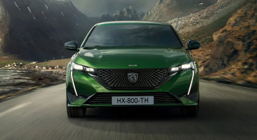 Zielony Peugeot 308 T7 jedzie drogą w malowniczym, górskim krajobrazie z widokiem na małą wioskę nad wodą.