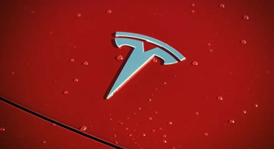 Błyszczące logo Tesli na czerwonej karoserii z kroplami deszczu. Czy Tesla jest tylko elektryczna?