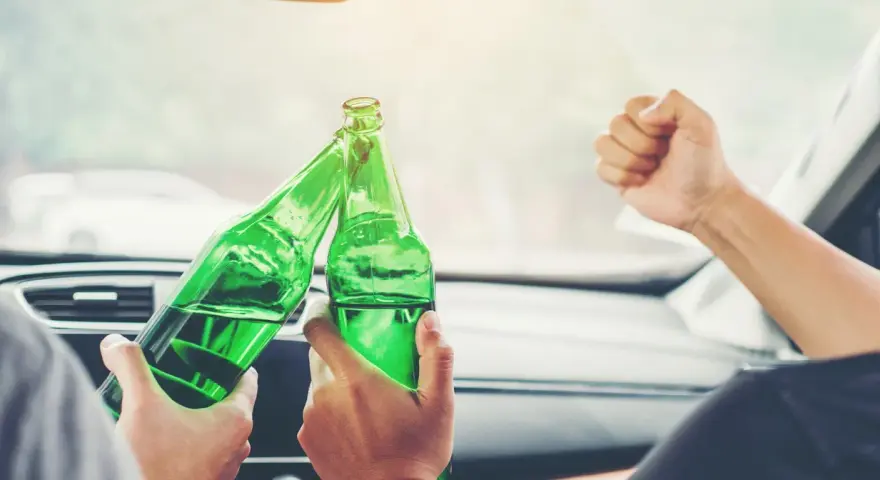 Dwie osoby w samochodzie wznoszą toast zielonymi butelkami. To nie jest dopuszczalna ilość alkoholu podczas jazdy.