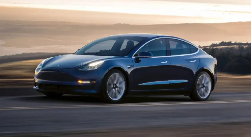 Niebieska Tesla pędzi po drodze, osiągając prędkość do 100 km/h. Tło rozmywa się w złotej poświacie zachodzącego słońca.