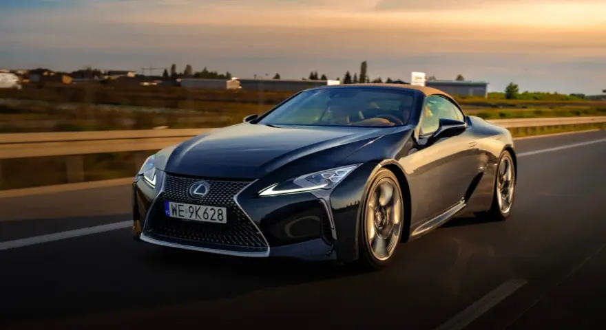 Ciemny Lexus LC 500 z beżowym dachem sunie po drodze podczas zachodu słońca. Sprawdź jego lexus lc 500 cena.