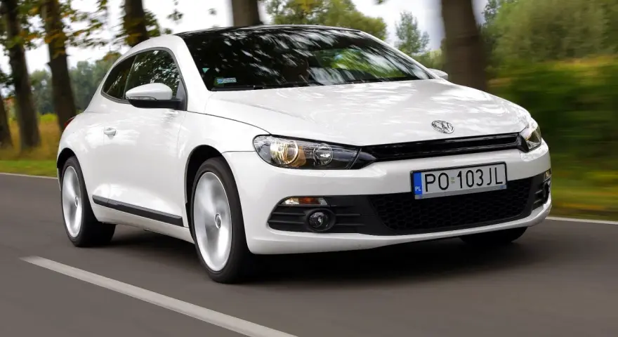 Biały Volkswagen Scirocco pędzi po drodze wśród drzew.