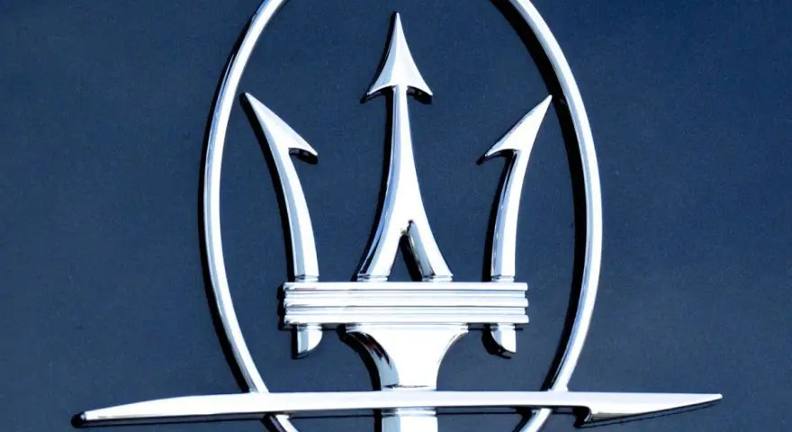 Błyszczący, chromowany trójząb Neptuna, czyli jak wygląda znaczek Maserati, na ciemnoniebieskim tle.