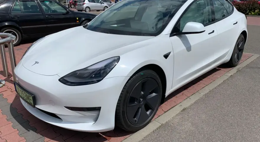 Biała Tesla Model 3 na parkingu przed sklepem. Zastanawiasz się, ile kosztuje najdroższa Tesla? Ten model to marzenie wielu kierowców.