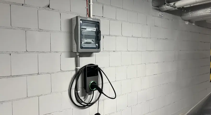 Nowoczesne gniazdo do ładowania samochodu elektrycznego Autel zamontowane na białej, ceglanej ścianie obok skrzynki z bezpiecznikami.