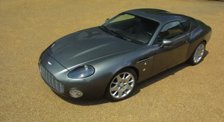 Szary Aston Martin DB9 na żwirowym podjeździe. Zastanawiasz się, ile kosztuje Aston Martin? To auto to marzenie wielu.