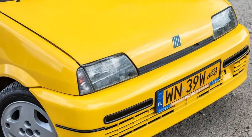Żółte tablice rejestracyjne na błyszczącym, żółtym samochodzie Fiat. Widoczny fragment maski, reflektor i zderzak.