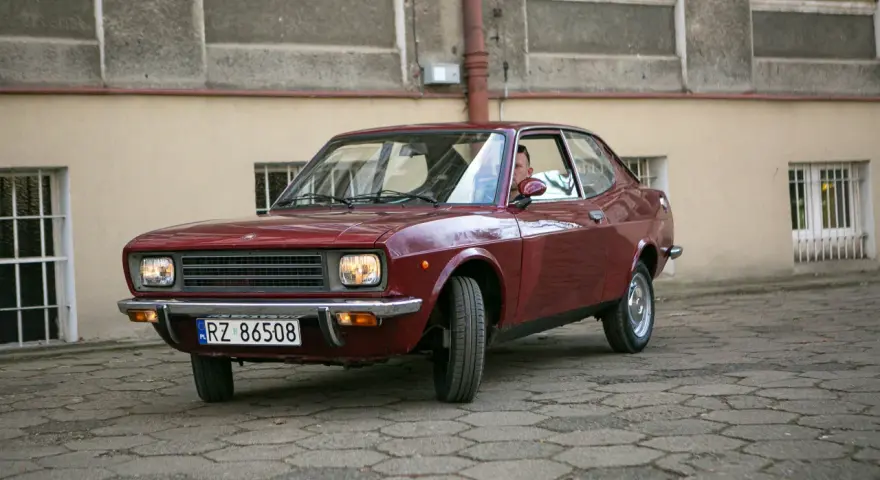 Czerwony fiat 128, klasyk z lat 70., zaparkowany na brukowanym dziedzińcu przed budynkiem.