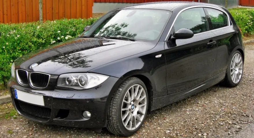 Czarny BMW e81, lśniący po deszczu, stoi na żwirowej drodze obok zielonego żywopłotu i drewnianego ogrodzenia.