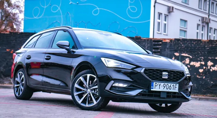 Czarny Seat Leon Sportstourer z polskimi tablicami rejestracyjnymi stoi na brukowanym placu przed budynkiem z niebieską, ozdobną ścianą.
