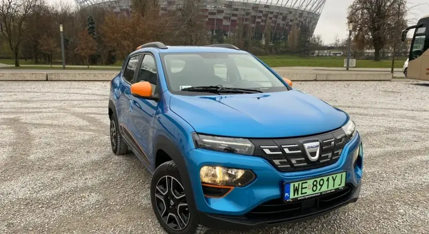 Niebieski Dacia Spring z pomarańczowymi akcentami na tle Stadionu Narodowego. Sprawdź atrakcyjną dacia spring cena!