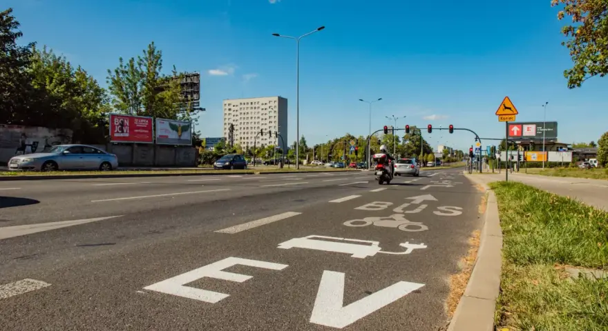 Na drodze oznaczenie "EV BUS" sugeruje, czy samochody elektryczne mogą jeździć buspasem. W tle budynek i ruch uliczny.