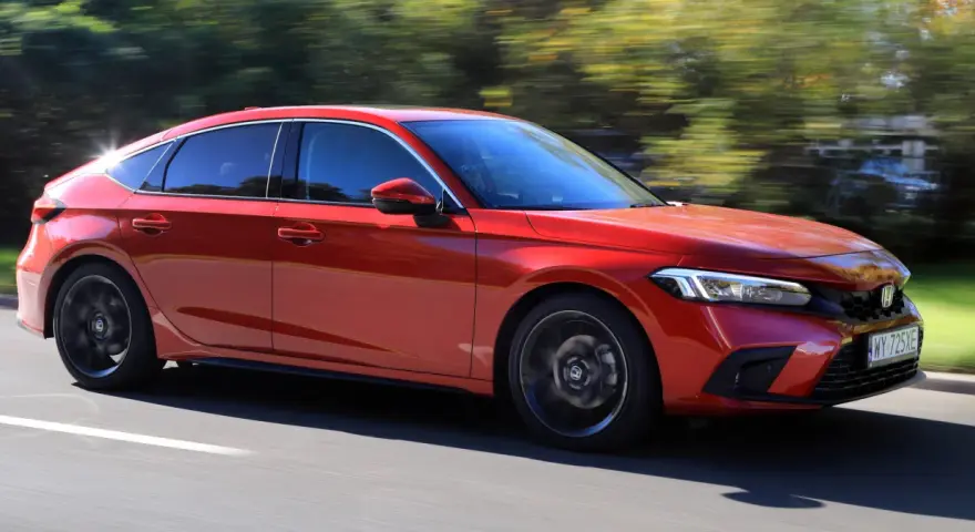 Czerwony Honda Civic 11. generacji pędzi po drodze, otoczony zielenią drzew.