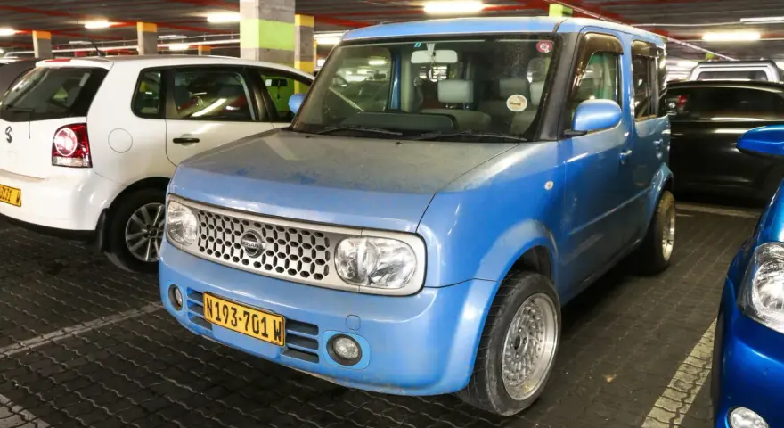 Niebieski Nissan Cube z charakterystycznym grillem i tablicą rejestracyjną N193-701 W, zaparkowany obok białego Volkswagena.