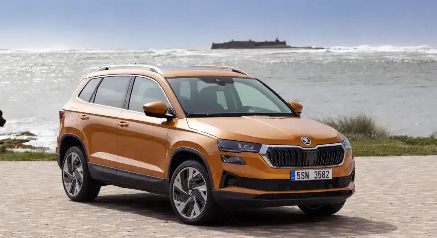 Pomarańczowa Skoda Karoq na tle morza i zamku. Sprawdź aktualny skoda cennik i wybierz swój wymarzony model.