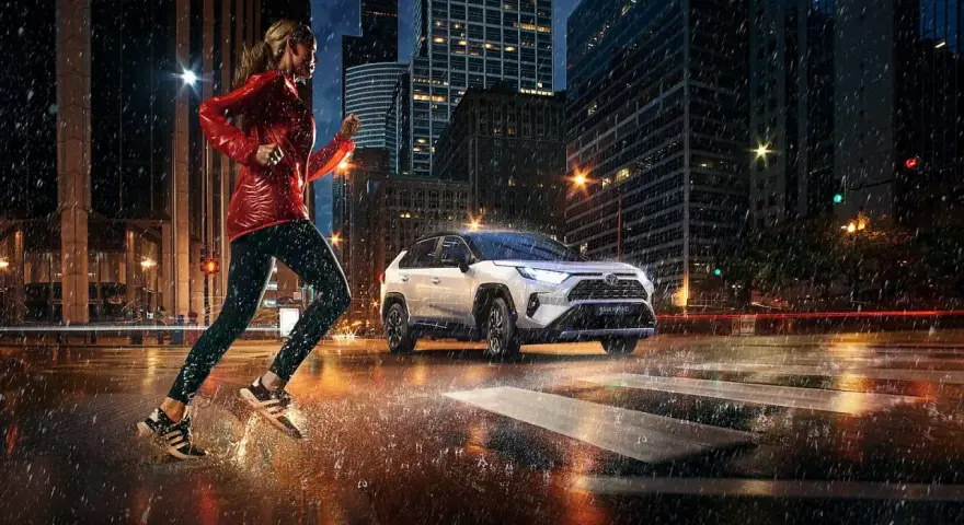 Biegaczka w deszczowy wieczór w mieście, obok niej biała Toyota RAV4 Hybrid. Zastanawiasz się, ile pali Toyota RAV4 Hybrid?