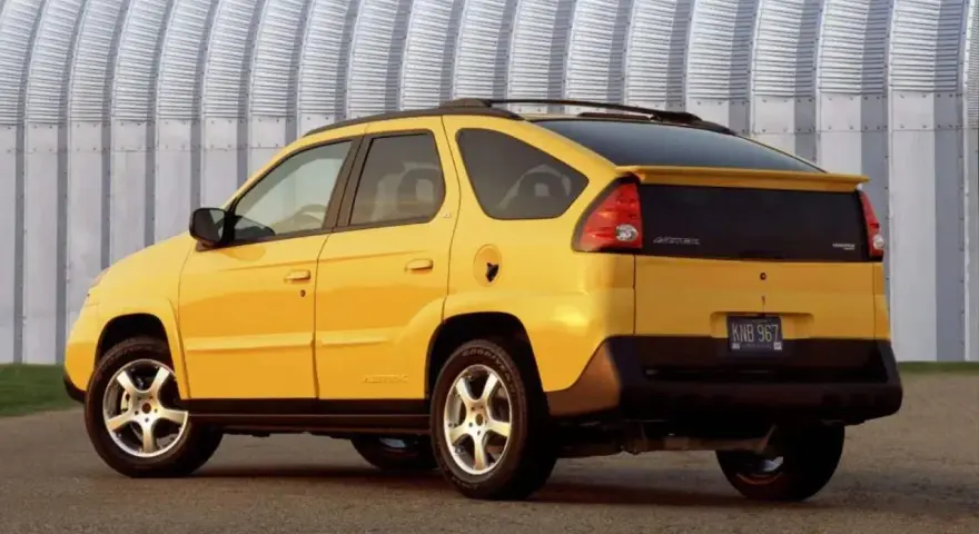 Żółty Pontiac Aztek z charakterystycznym tyłem i spojlerem, zaparkowany przed falistą, metalową ścianą.