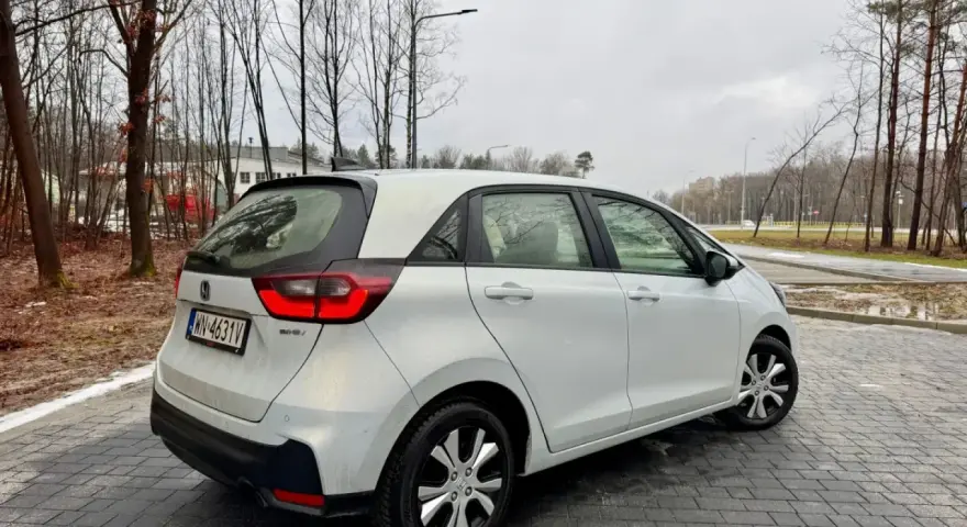 Biały Honda Fit stoi na mokrej, brukowanej nawierzchni. W tle drzewa i budynki.