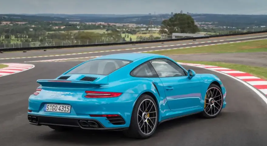 Błękitne Porsche 911 Turbo S na torze wyścigowym, z widokiem na miasto w tle.