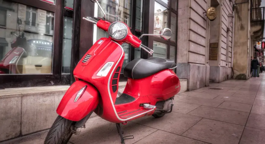 Czerwony skuter Vespa, idealny do zdobycia prawa jazdy AM, stoi na chodniku przed budynkiem.
