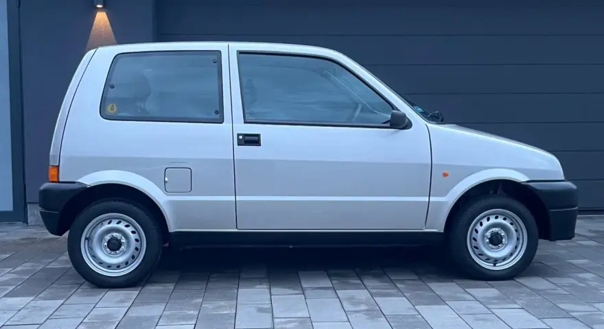 Srebrny Fiat Cinquecento na tle ciemnego garażu, z charakterystycznymi felgami i czarnymi zderzakami.