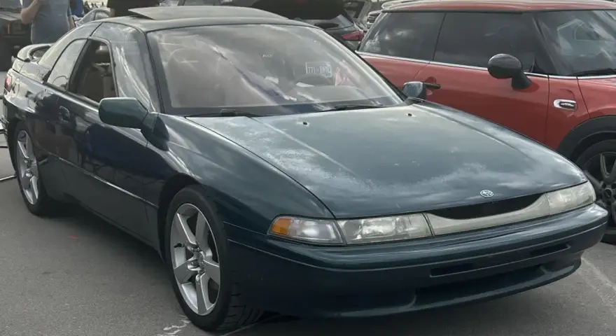 Subaru SVX w ciemnozielonym kolorze, z charakterystycznymi oknami w stylu "glass-canopy", zaparkowany obok czerwonego samochodu.