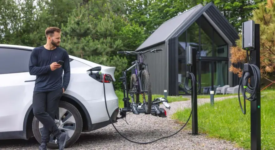 Mężczyzna z telefonem przy białym samochodzie elektrycznym, który ładuje się obok stojaka z rowerami. Obok stoją dwie ładowarki do samochodów elektrycznych.