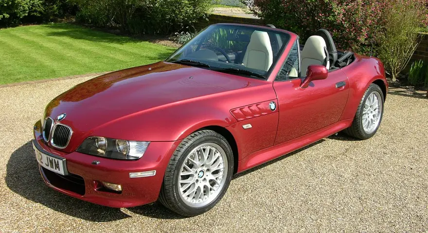 Czerwony kabriolet BMW E37 z jasnym wnętrzem zaparkowany na żwirowej nawierzchni, w tle zielony trawnik i kwitnące krzewy.