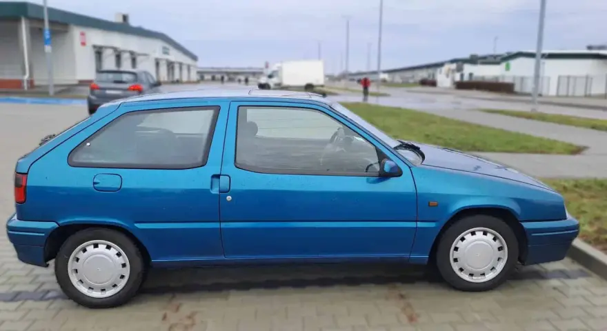Niebieski Citroen ZX stoi na parkingu. W tle widać budynki i białą ciężarówkę.