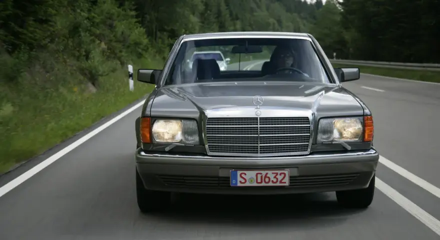 Szary Mercedes W126 jedzie drogą wśród zielonych lasów.