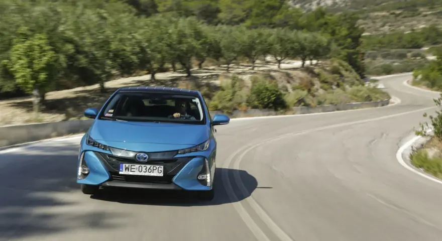 Niebieska Toyota Prius pokonuje zakręt na malowniczej drodze. Zastanawiasz się, ile pali Toyota Prius? Ten model jest znany z oszczędności.