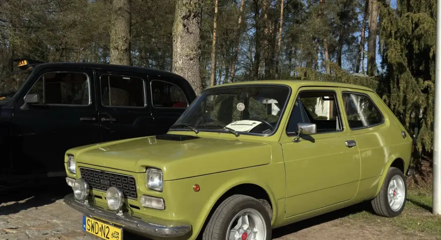 Zielony fiat 127 z lat 70. stoi obok czarnej taksówki na tle drzew.