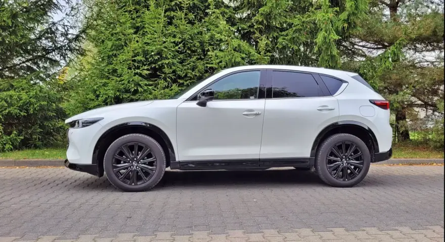 Nowa Mazda CX-5 w białym kolorze, zaparkowana na brukowanym podjeździe, otoczona zielenią drzew.