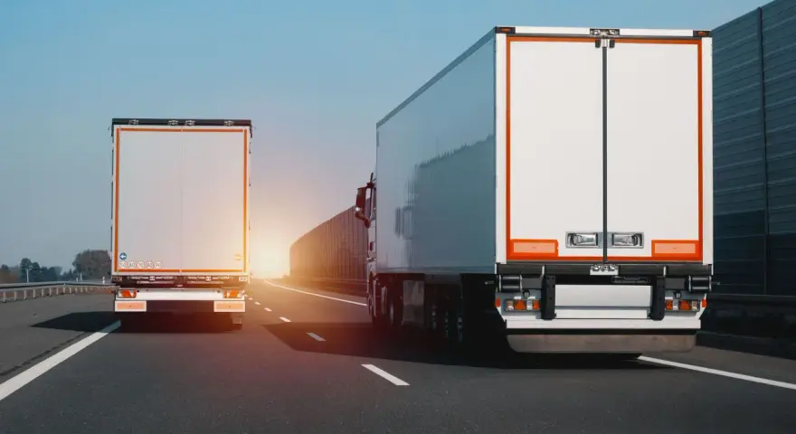 Dwa białe tiry jadą autostradą. Na jednym z nich widać znak zakazu wyprzedzania dla ciężarówek.