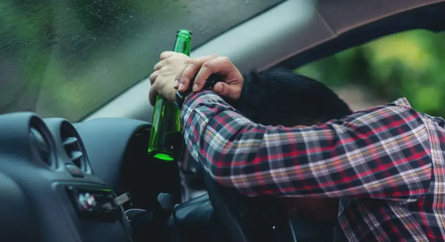 Mężczyzna w kratę trzyma butelkę, opierając głowę na kierownicy. To obraz ostrzegawczy przed jazdą pod wpływem alkoholu, przypominający o zagrożeniach i potrzebie blokady alkoholowej.