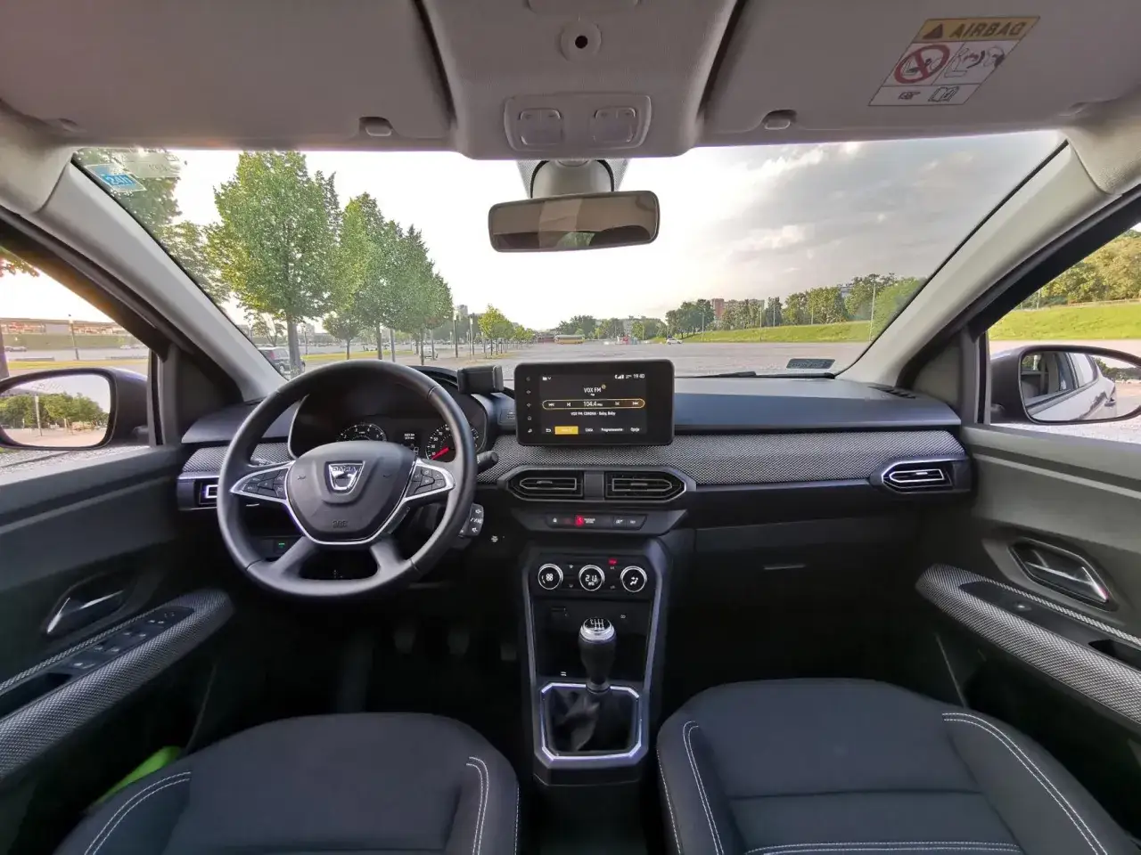 Wnętrze Dacia Sandero z ekranem multimedialnym, kierownicą i fotelami. Za oknem drzewa i parking.
