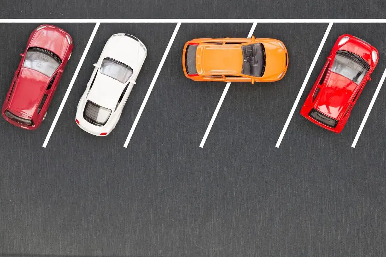 Czerwony, biały i pomarańczowy samochodzik na ciemnym tle, z białymi liniami wyznaczającymi miejsca parkingowe. Idealny przykład parkowania równoległego tyłem.