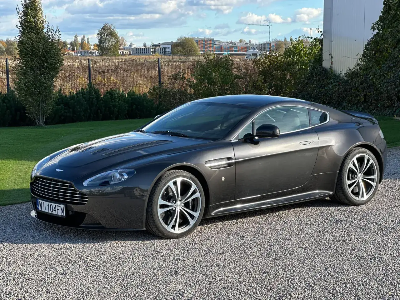 Elegancki, ciemnoszary Aston Martin V8 Vantage na żwirowym podjeździe. Zastanawiasz się, ile kosztuje Aston Martin?