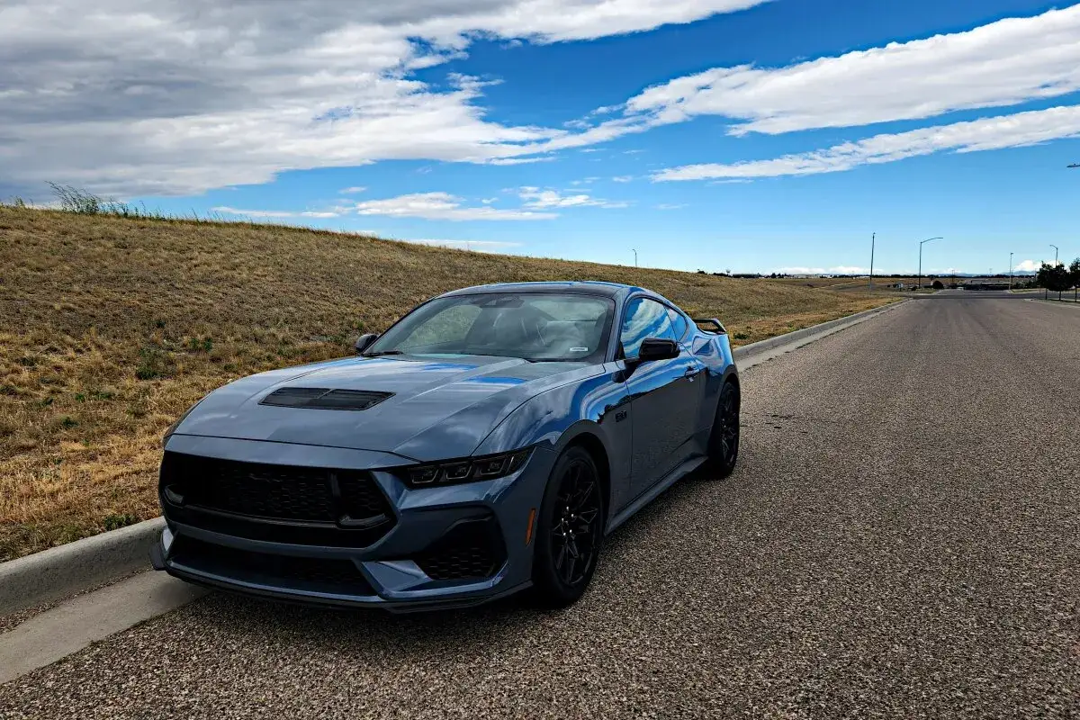Szary Mustang GT 5.0 z niebieskimi pasami na masce stoi na poboczu drogi, pod błękitnym niebem z chmurami.