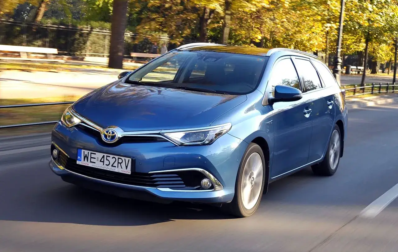 Niebieska Toyota Auris Hybrid w wersji kombi jedzie po drodze, a jesienne drzewa tworzą tło. Zastanawiasz się, ile pali Toyota Auris Hybrid?