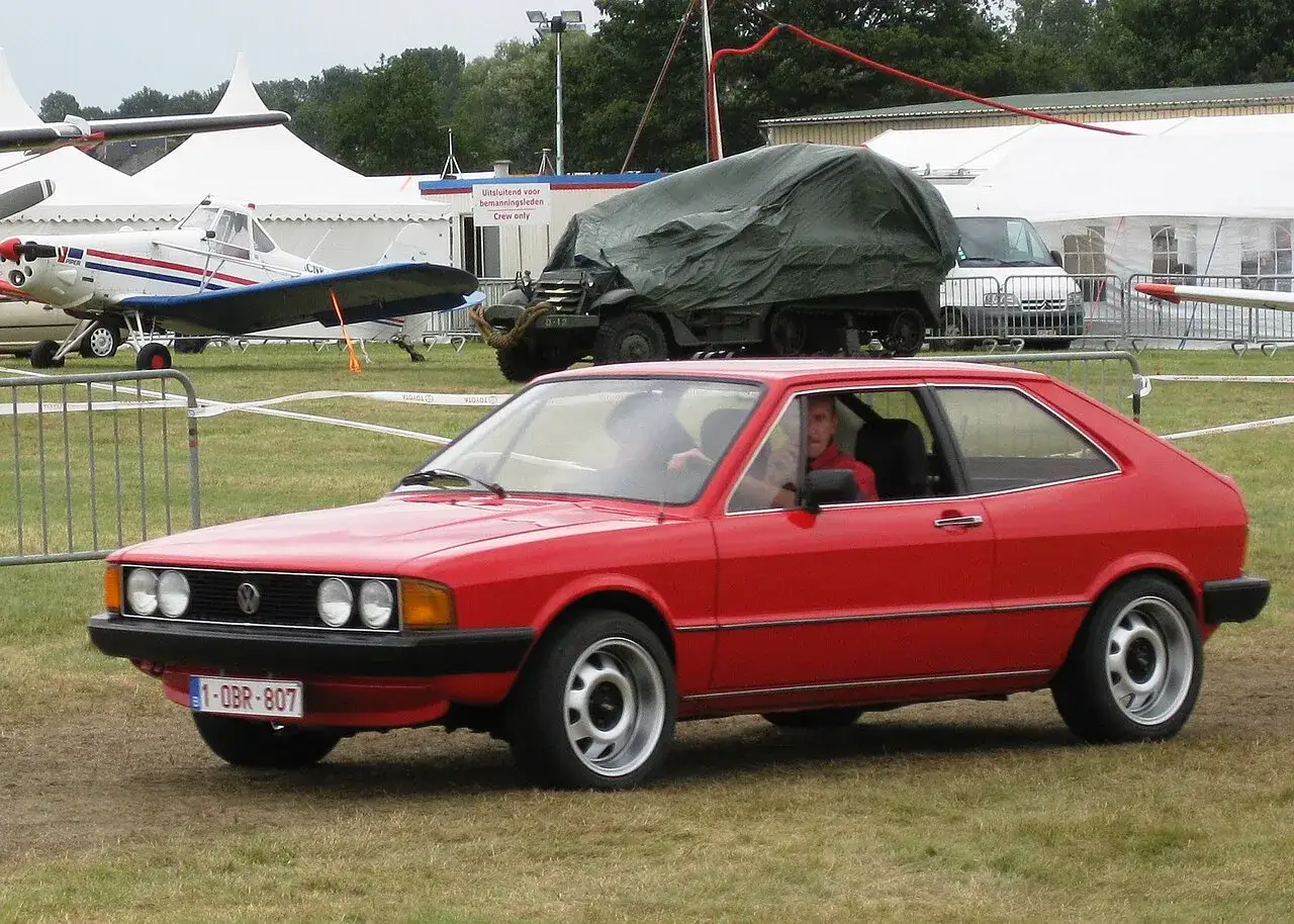 Czerwony Volkswagen Scirocco na trawiastym polu, obok samolotu i zakrytej ciężarówki.