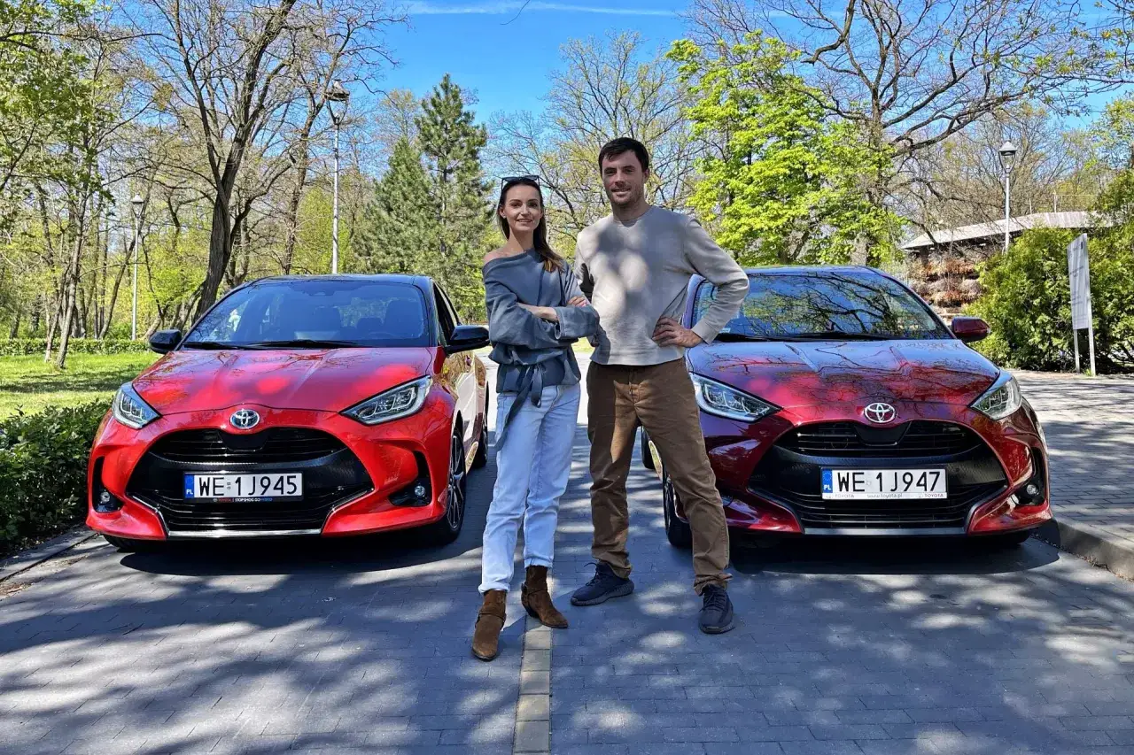Para testuje, ile pali Toyota Yaris Hybrid. Dwa czerwone Yarisy hybrydowe stoją obok siebie, a na ich tle para młodych ludzi.