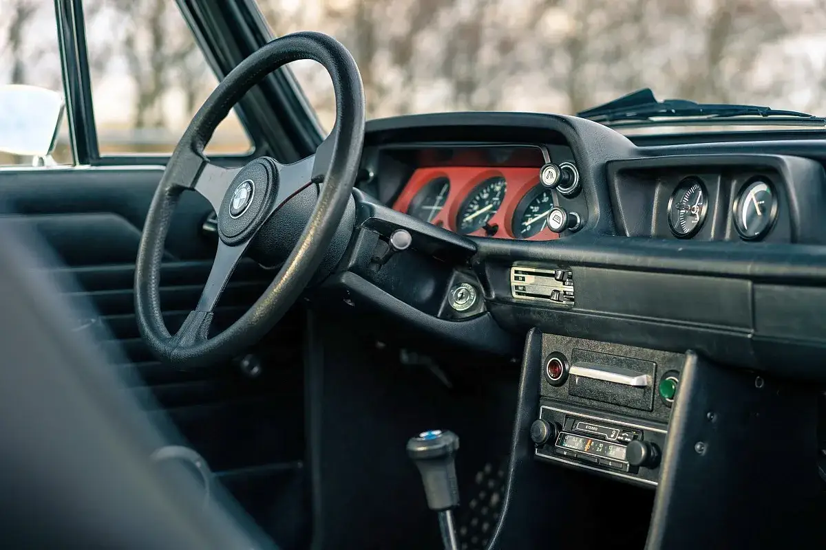 Wnętrze klasycznego BMW E20 z czarną kierownicą, czerwonymi tarczami zegarów i radiem.